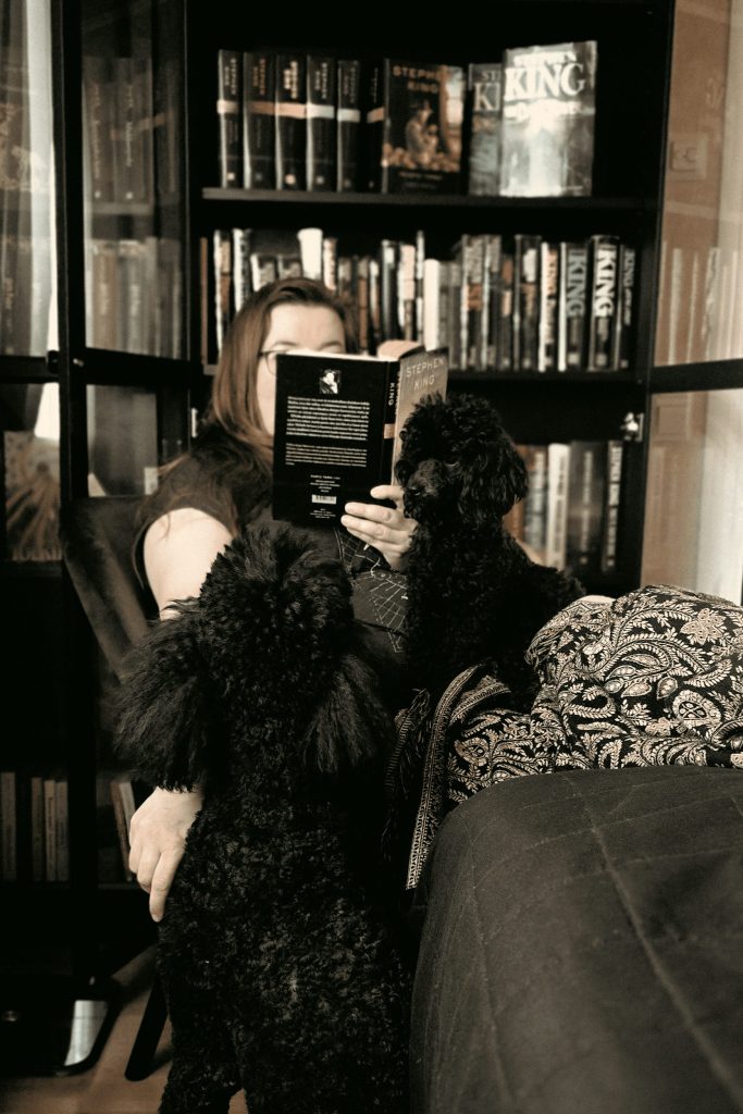 Juontaja Marin kuva. Kuvassa Mari istuu kirjahyllyn edessä lukemassa Mustan Tornin ensimmäistä osaa Revolverimiestä. Pieni villakoira Turo istuu Marin sylissä ja Isovillakoira Ippe vieressä. Kirjahylly on täynnä Stephen Kingin kirjoja